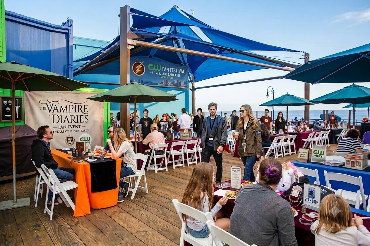 Fans, autograph tables and shaded seating on a pier boardwalk beneath a CW Fan Festival banner, with the ocean behind